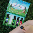 A person lying on grass holds a small essential oil roller bottle next to an open illustrated gift set containing four oil bottles and a white tube. The set lid reads 'Feel Better with Nature's Magic' and shows a colorful forest illustration.