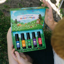 A person outdoors holds an open gift box containing five small essential oil bottles. The box lid has colorful cartoon artwork with the text 'Feel Better with Nature's Magic.