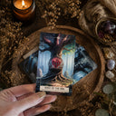 A hand holds a tarot card titled 'Root of the Oak' in a styled arrangement with dried flowers, crystals, a candle, and another card on a wooden tray.