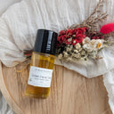 Facial oil bottle with a black cap on a wooden tray next to dried flowers, all on a white fabric background.