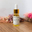 Bottle of Wildroot Alchemy Sylvan Glow oil on a wooden surface with flowers in the background