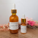 Two bottles of 'Lover's Aura' by Widroot Alchemy on a wooden surface with flowers in the background.