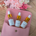 Three dropper bottles with labels on a pink pouch, with pink flowers in the background.