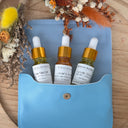 Three small bottles with gold caps and white labels on a blue pouch with dried flowers around.