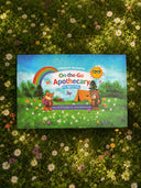 A children's activity book titled 'On the Go Apothecary' by Wildroot Alchemy lies flat on a bed of green grass and small white daisies. The cover features a colorful cartoon scene with a rainbow, campfire, tent, sun, and two animals.