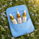 A light blue leather pouch with three small dropper bottles of oil in it resting on a bed of green foliage and small white flowers.